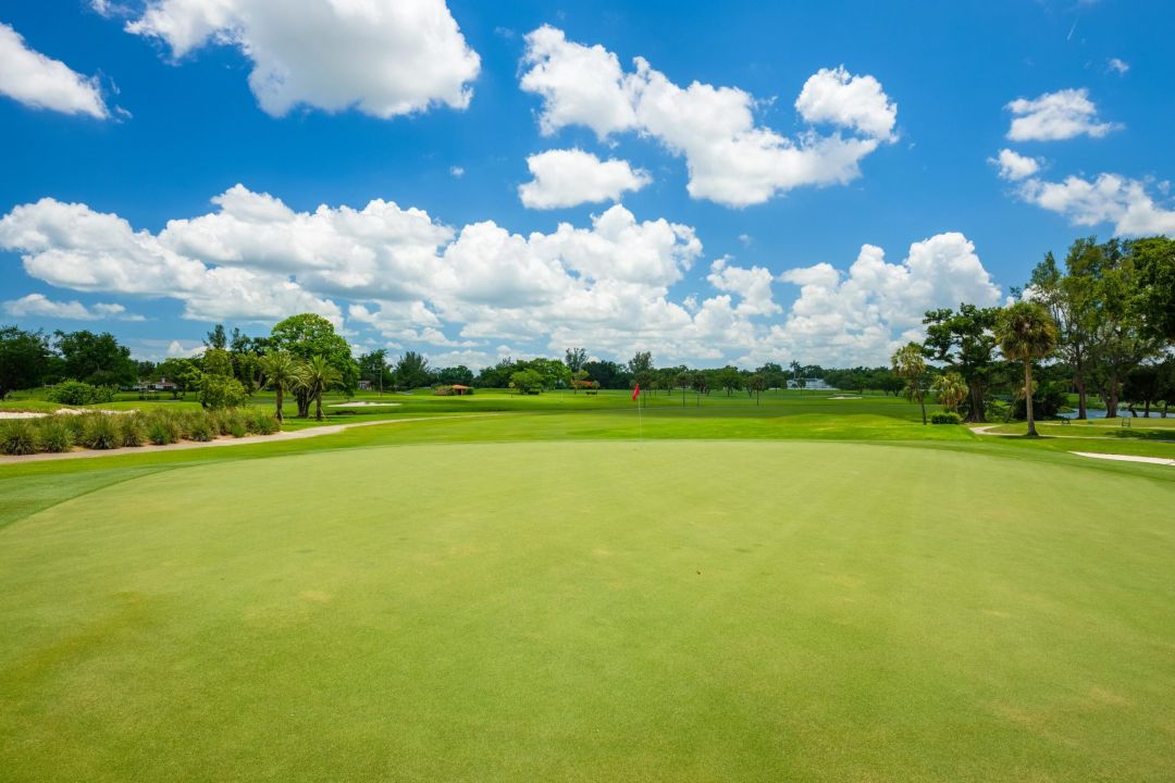 Inspira Naples A bright, sunny day on a golf course with green grass, trees, and a blue sky with scattered clouds.