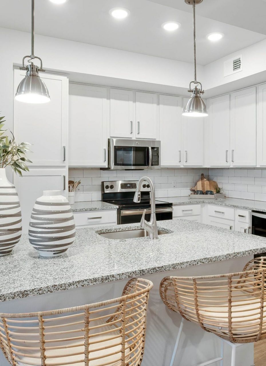Inspira Naples Modern white kitchen with granite island, wicker bar stools, stainless steel appliances, and pendant lights.