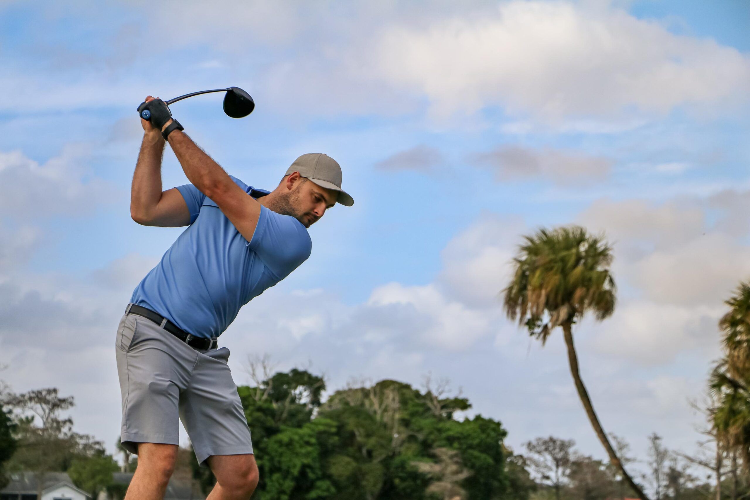 Inspira Naples Man in blue shirt and cap swinging a golf club outdoors, with trees and a cloudy sky in the background.