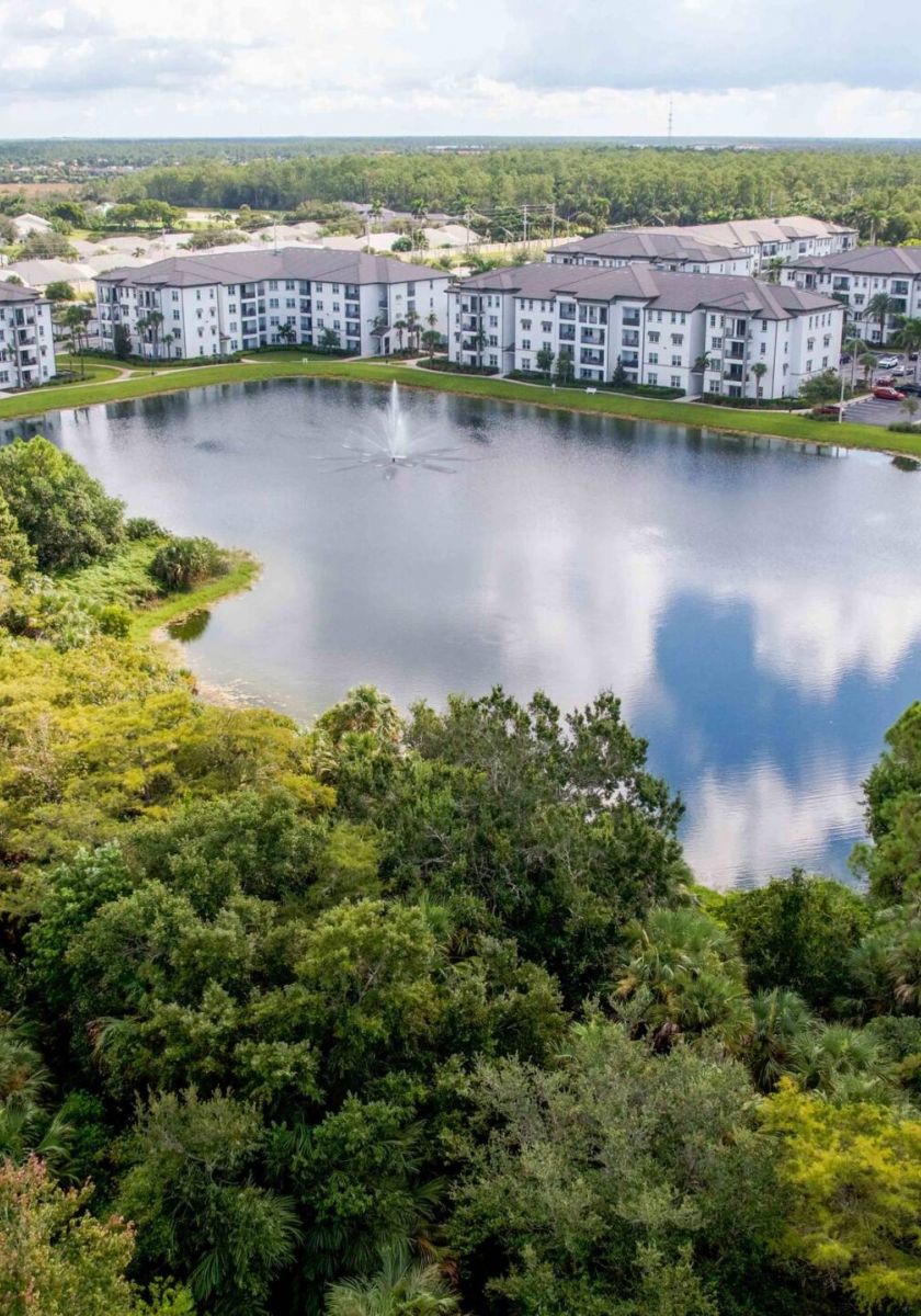 Inspira Naples Aerial view of apartments surrounding a pond with trees and a road nearby under a cloudy sky.