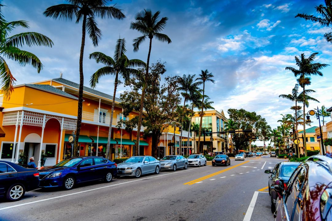 Inspira Naples A vibrant street lined with palm trees, colorful buildings, and parked cars under a partly cloudy sky.