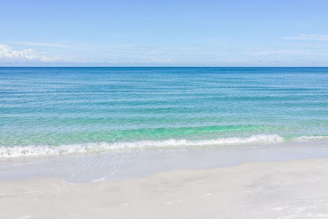 Inspira Naples Calm turquoise waves gently washing onto a bright sandy beach under a clear blue sky.
