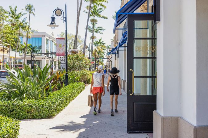 Inspira Naples Two people walk down a sunny, palm-lined sidewalk with shopping bags beside storefronts.