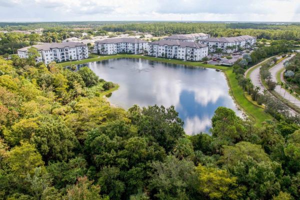 Inspira Naples Aerial view of apartments surrounding a pond with trees and a road nearby under a cloudy sky.
