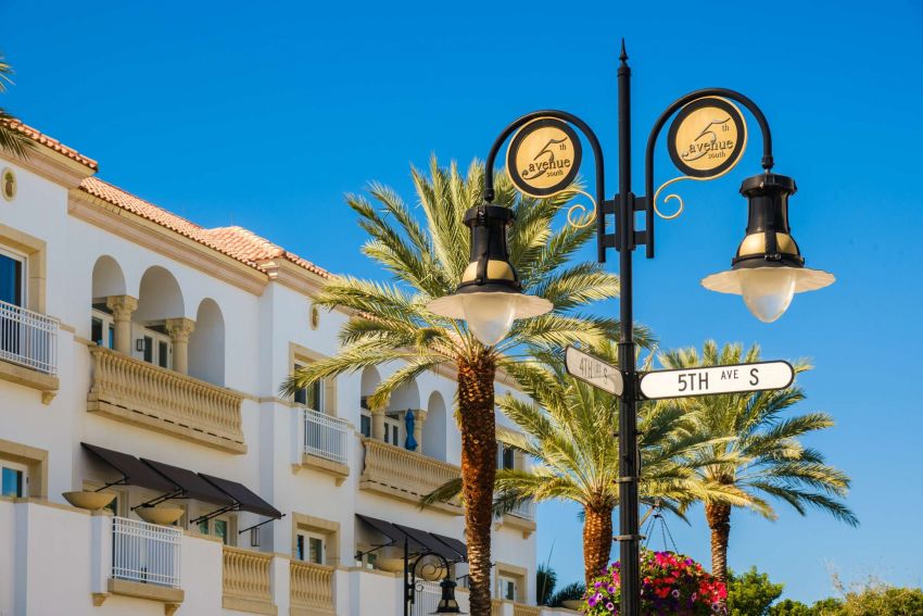 Inspira Naples Streetlamp and 5th Ave S sign in front of a white building with palm trees under a clear blue sky.