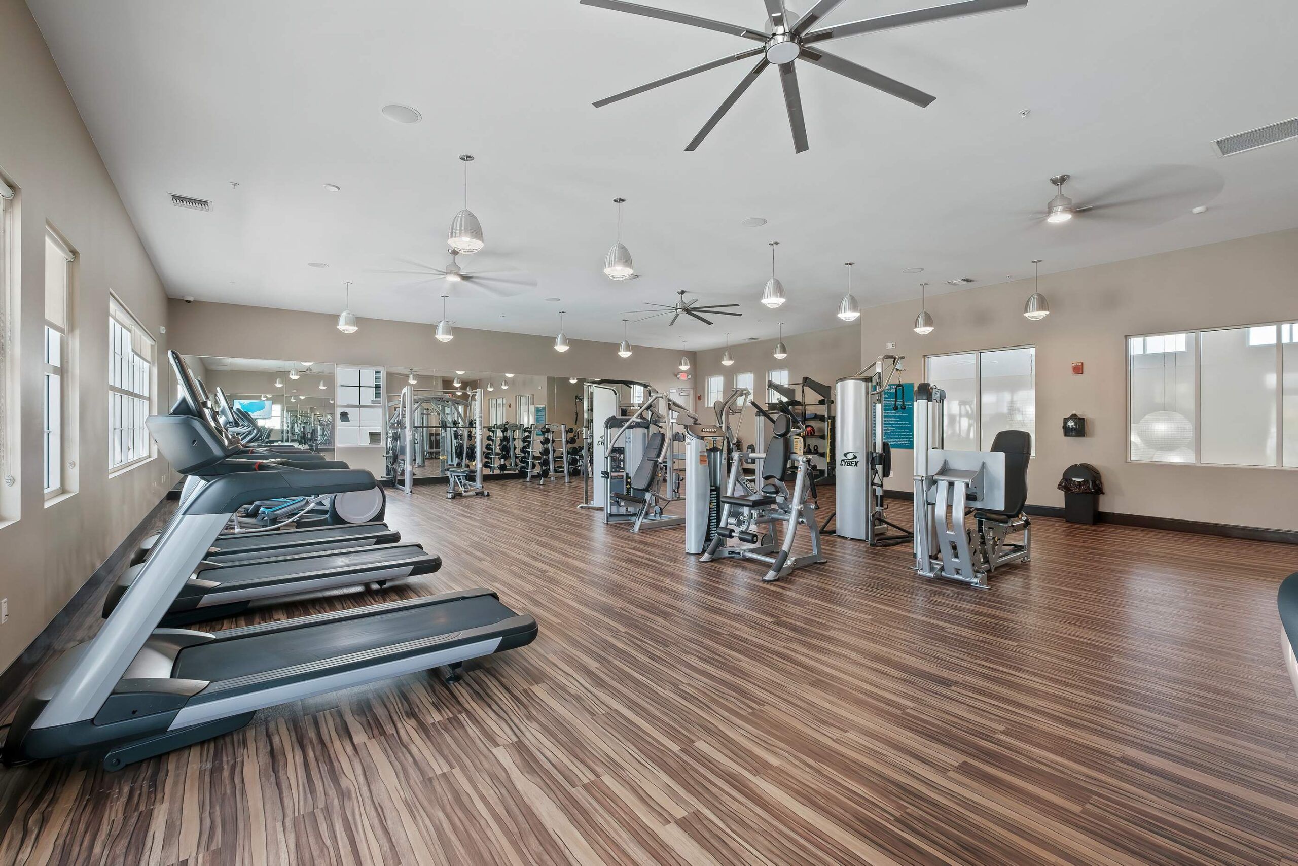 Spacious Inspira Naples gym with treadmills, exercise machines, wood flooring, large windows, and ceiling fans.
