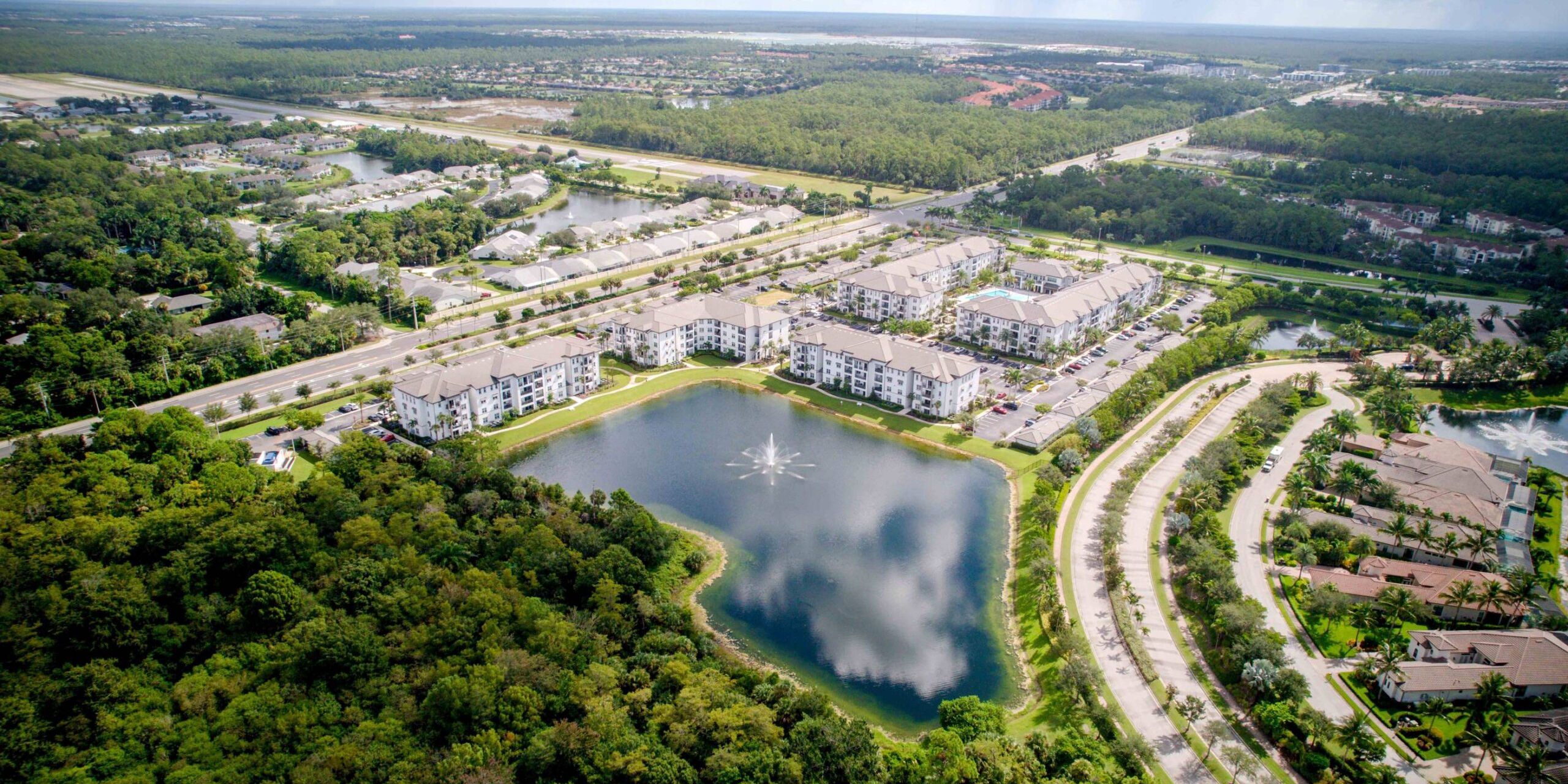 Inspira Naples Aerial view of apartment buildings around a pond with surrounding trees and roads.
